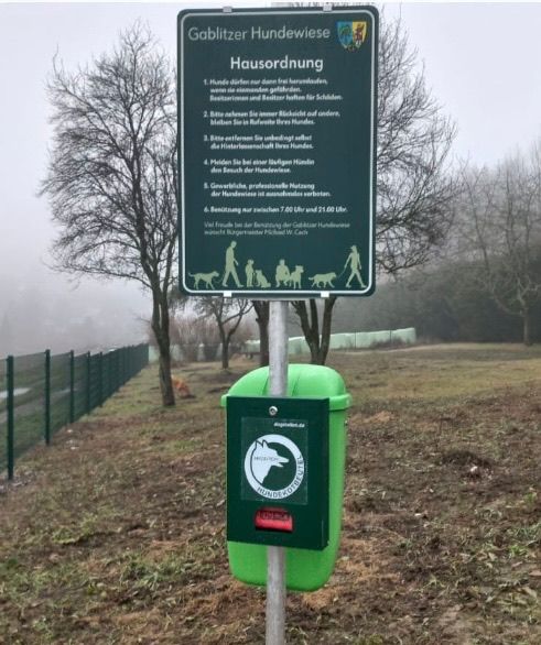 Grüne Tafel mit deutschem Text steht neben einem grünen Mülleimer. Die Tafel gibt Richtlinien und Regeln für die Entsorgung von Hundekot vor.