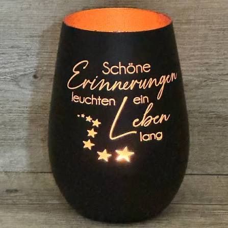 Ein schwarzer zylindrischer Kerzenhalter mit goldenem Text, der 'Schöne Erinnerungen leuchten ein Leben lang' und Sternen sagt, auf einer Holzoberfläche.