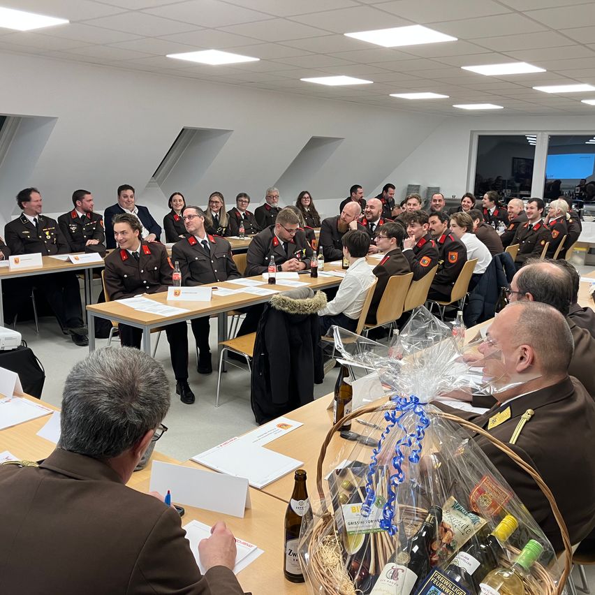 Eine Gruppe von Menschen in Uniform sitzt um Tische in einem Konferenzraum herum und bespricht etwas. Vor ihnen stehen Flaschen und ein Korb mit Wein.
