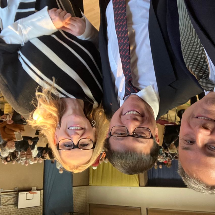 Drei Personen lächeln in die Kamera. Die Frau in der Mitte hat blonde Haare und trägt eine Brille. Die beiden Männer tragen Krawatten und Brillen. Sie scheinen sich bei einer Veranstaltung zu befinden.