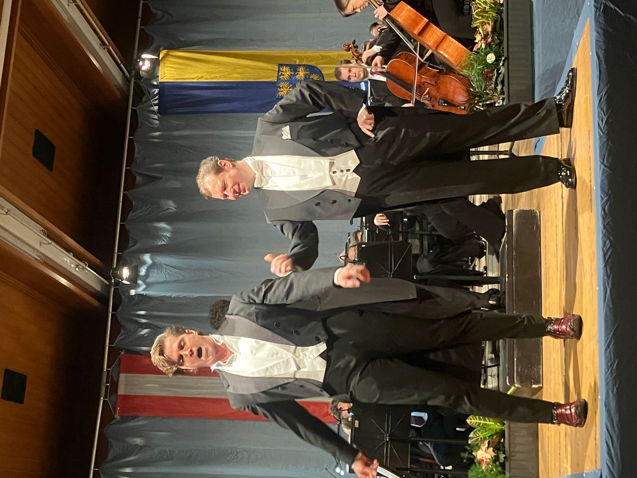 Zwei Männer in formellen Anzügen stehen auf der Bühne, möglicherweise bei einer Aufführung. Hinter ihnen spielen andere Celli und Violinen. Die Bühne ist mit rot-weiß-blauen Bannern und Holzboden dekoriert.