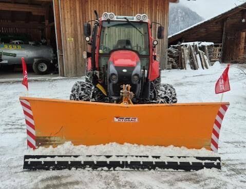 Ein roter Traktor mit einem Schneepflug-Aufsatz steht vor einem Holzgebäude in einem verschneiten Bereich.