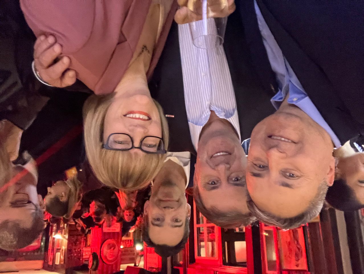 Eine Gruppe von Menschen posiert für ein Foto in einem schwach beleuchteten Raum. Eine Frau mit Brille und Halskette lächelt. Zwei Männer schauen zur Kamera. Rote Lichter beleuchten den Raum.