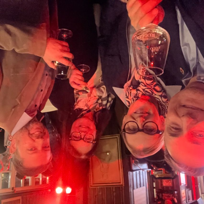 Vier Personen versammeln sich in einem schwach beleuchteten Raum und halten jeweils ein Glas. Zwei von ihnen tragen Brillen, einer lächelt. Rote Lichter und gerahmte Bilder schmücken den Hintergrund.