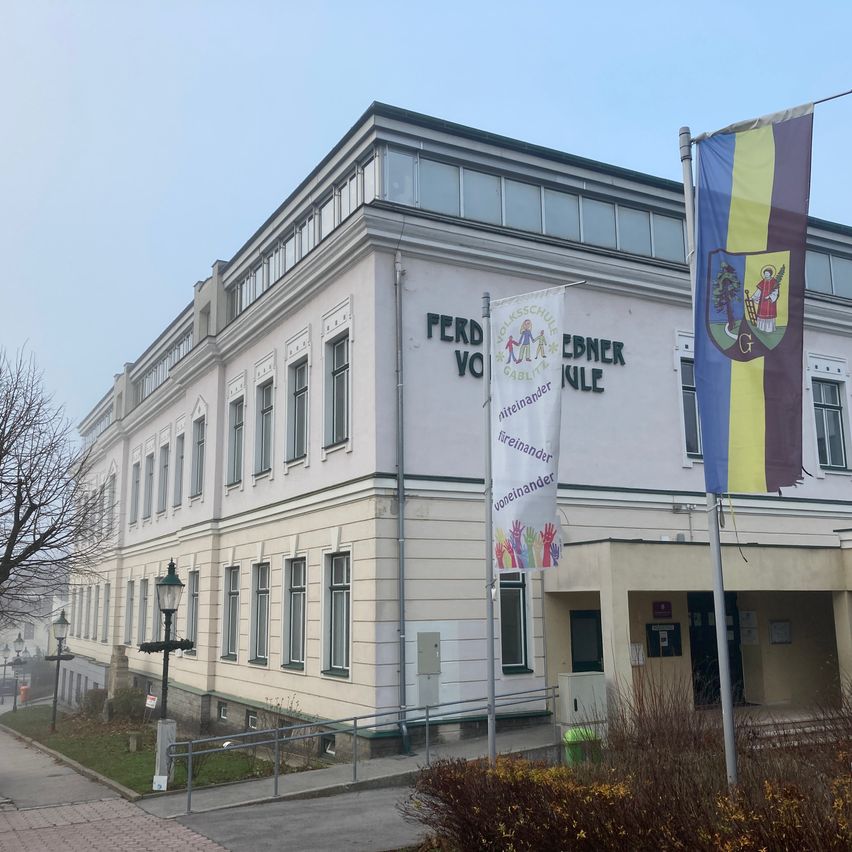 Ein großes weißes Gebäude mit vielen Fenstern, beschriftet mit 'Perd vo Ebdner Kle', steht an einer Straße. Vor dem Gebäude weht eine Fahne mit einem Wappen.