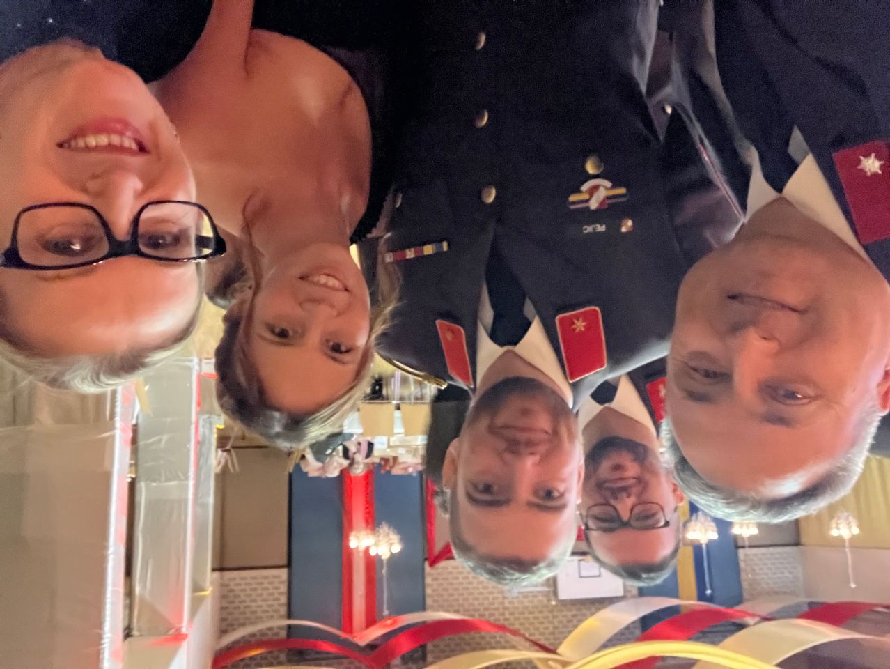 Eine Gruppe von Menschen in formeller Kleidung, möglicherweise bei einer Feier, posiert für ein Spiegel-Selfie. Ein Mann trägt eine Brille. Der Hintergrund zeigt Dekorationen und eine Kronleuchter.