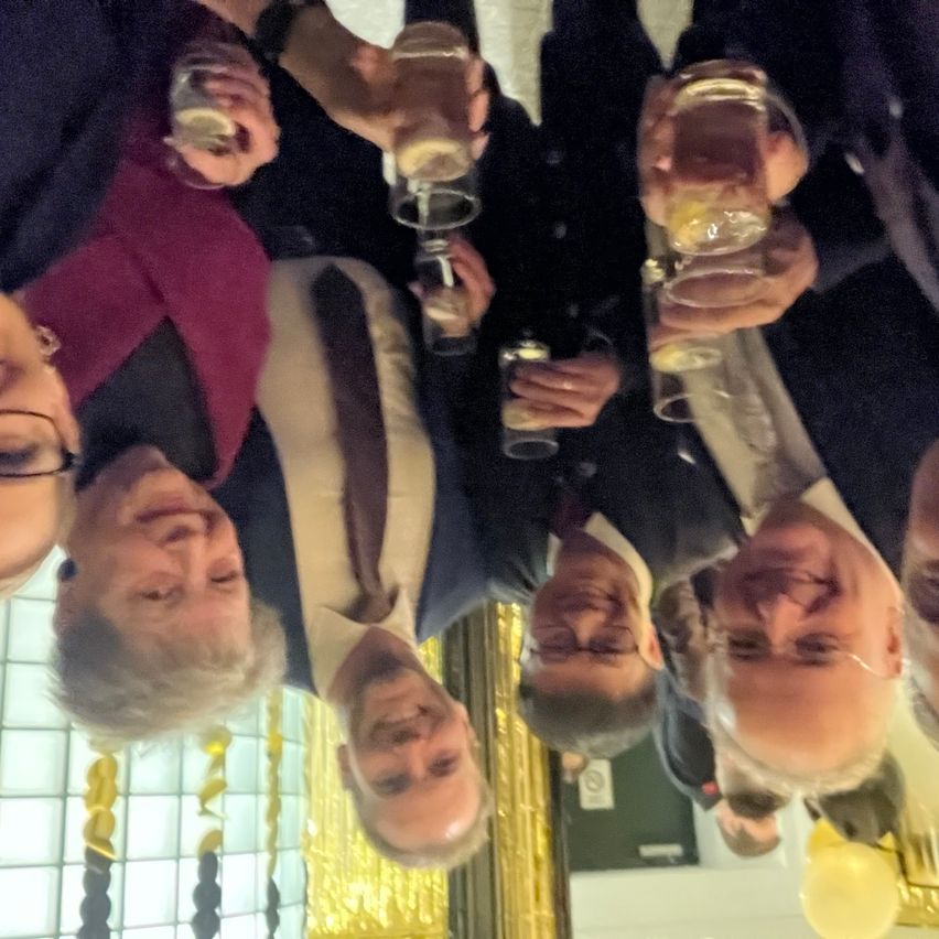 Eine Gruppe von Menschen in formeller Kleidung hält Gläser mit Getränken hoch. Das Bild ist aus einer niedrigen Perspektive aufgenommen und zeigt ihre Gesichter und einige Dekorationen im Hintergrund.