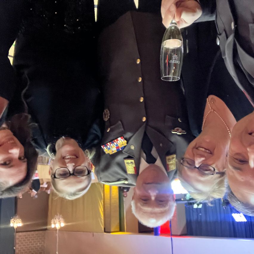 Fünf Personen stehen zusammen, lächeln und posieren für ein Foto. Ein Mann hält eine Flasche. Drei von ihnen tragen Brillen. Zwei sind in Militäruniformen. Im Hintergrund befindet sich ein blauer Vorhang und Lichter.