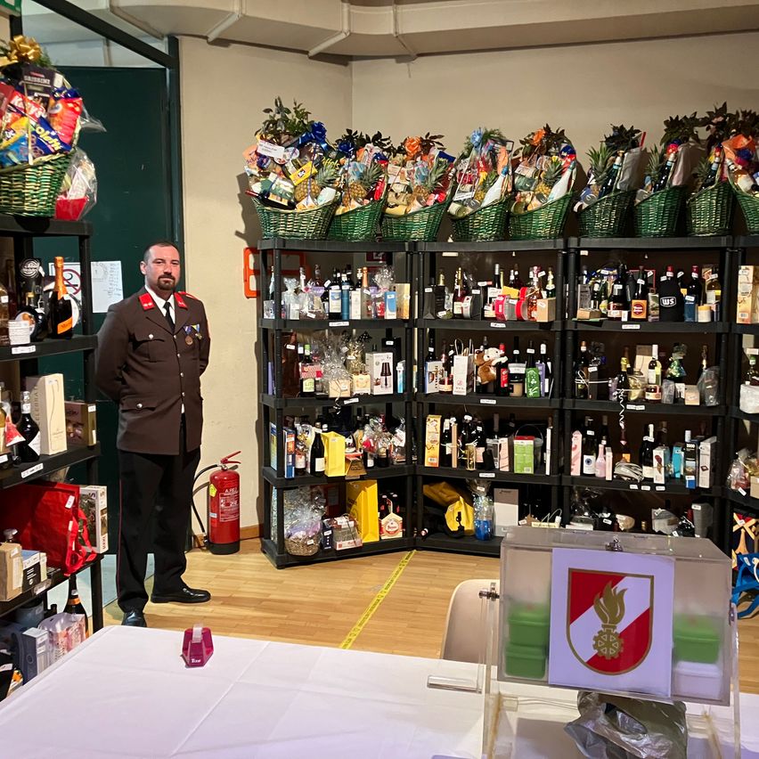Ein Mann in Uniform steht in einem Raum mit Regalen, die mit verschiedenen Geschenkkörben und Flaschen gefüllt sind, einem Feuerlöscher in der Nähe und einem Schreibtisch mit einer Box vor ihm.