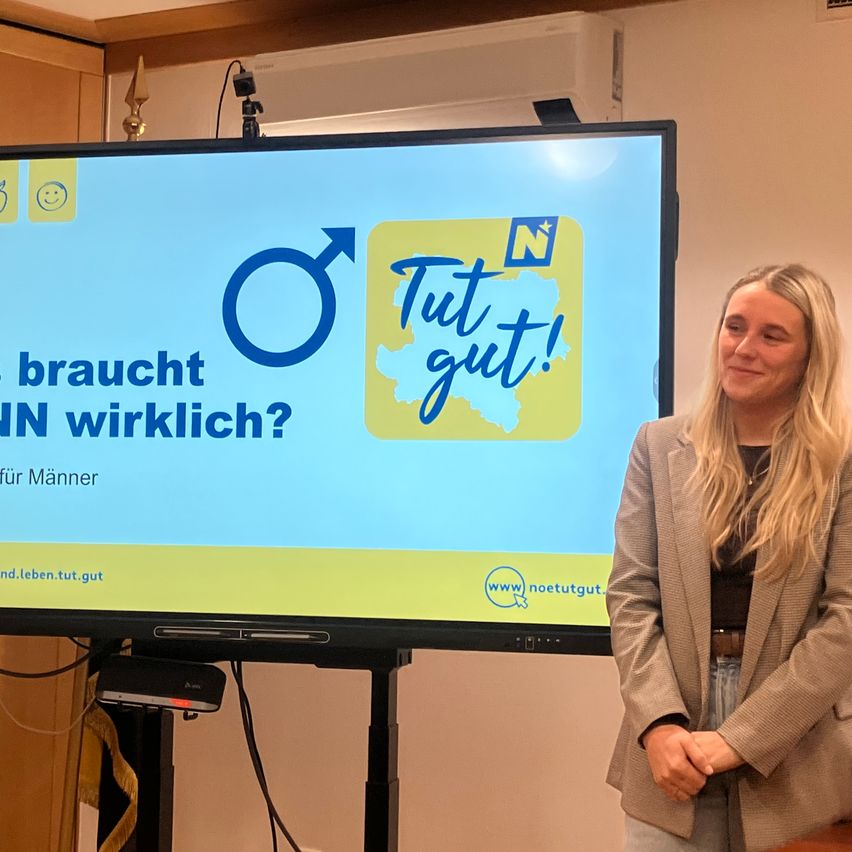 Eine Frau steht vor einem Bildschirm, der eine Präsentation zeigt. Der Bildschirm trägt die Aufschrift 'Tut gut! Braucht NN wirklich?' mit einer Karte und einem männlichen Symbol. Die Frau lächelt.