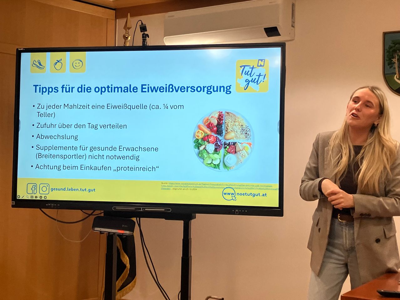 Eine Frau steht neben einer Projektionsleinwand mit einer Präsentation über Proteinaufnahme. Die Leinwand zeigt einen Teller mit Essen und Text über optimale Proteinversorgung.