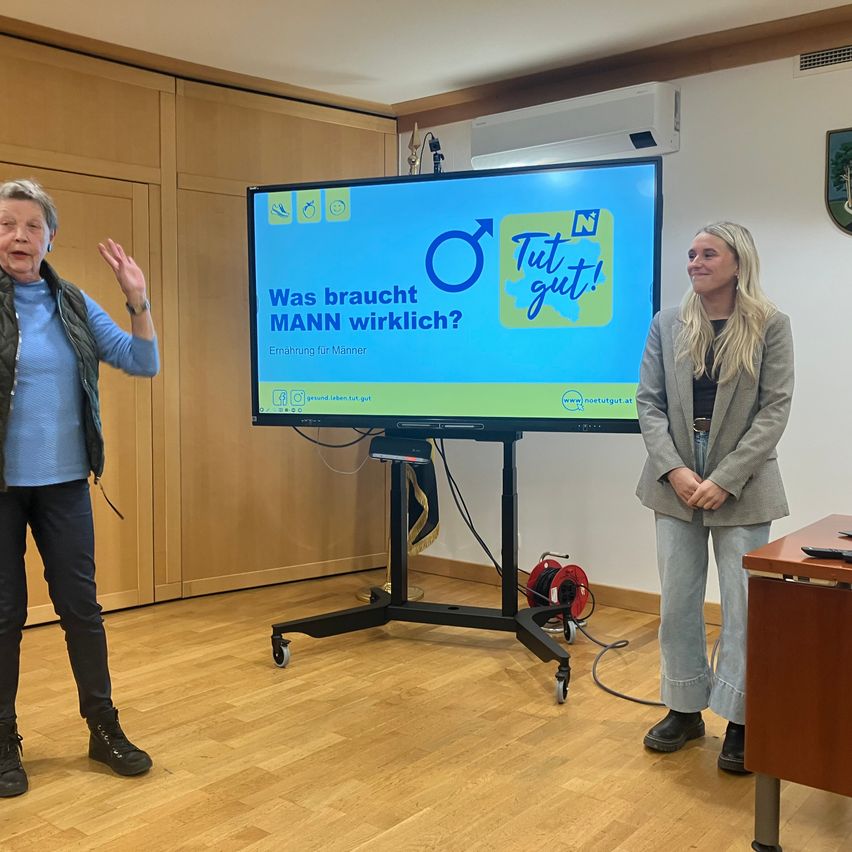Zwei Frauen stehen vor einem Monitor mit einer Bildschirmanzeige einer Kampagnenbotschaft über männliche Ernährung. Die Botschaft lautet 'Was braucht ein Mann wirklich?' Die Frau auf der linken Seite gestikuliert, während sie spricht.
