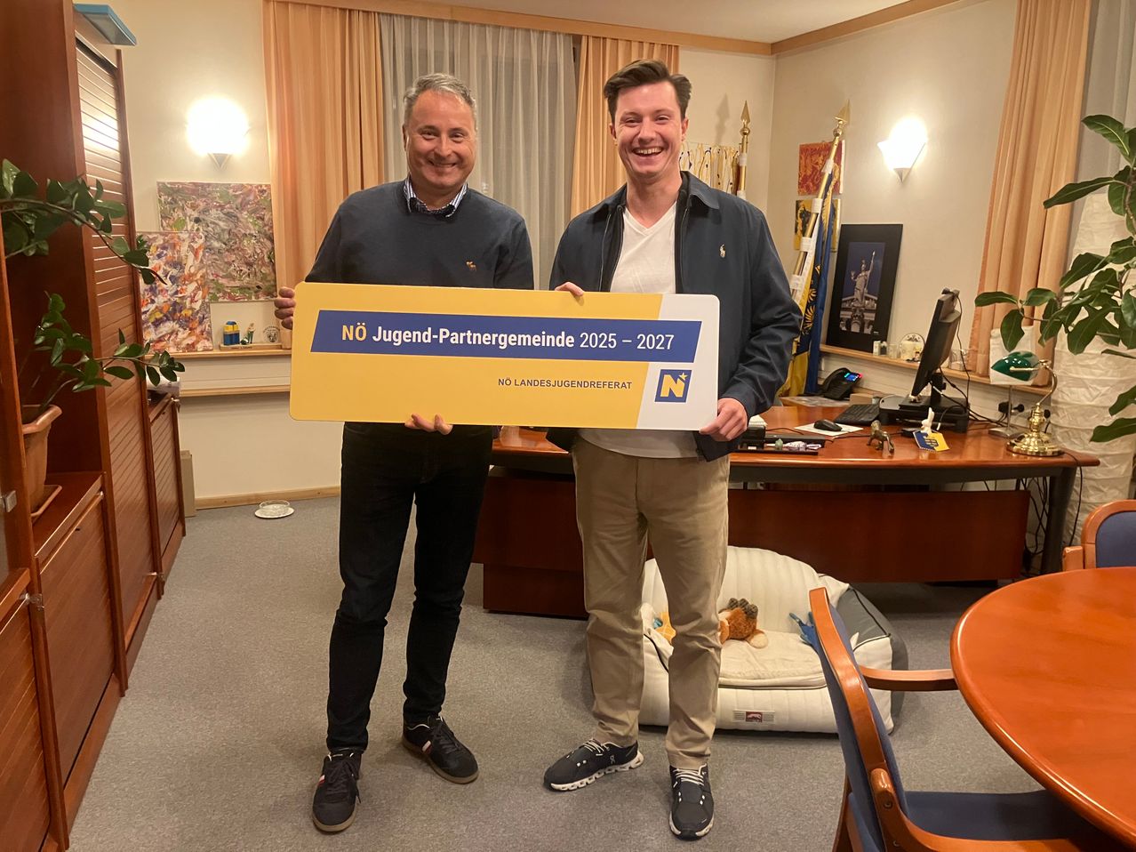 Zwei Männer halten ein großes gelbes Banner in einem Raum. Sie lächeln und einer hält das Banner mit beiden Händen. Hinter ihnen steht ein Schreibtisch mit Computer, Telefon und anderen Gegenständen. Ein Hundebett steht auf dem Boden.