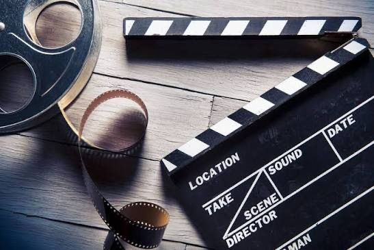 Ein Filmklapper, eine Filmrolle und ein Filmstreifen liegen auf einer Holzoberfläche. Der Filmklapper hat Bezeichnungen wie 'Location', 'Szene', 'Regisseur' und 'Ton'. Die Filmrolle hat Löcher und Zahnräder. Der Filmstreifen hat Zahnräder.