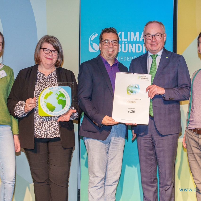 Five people stand together holding awards. Two men hold a certificate. A woman holds an award with a globe on it. They are standing in front of a screen with the text 'KLIMA BÜNDNIS'.