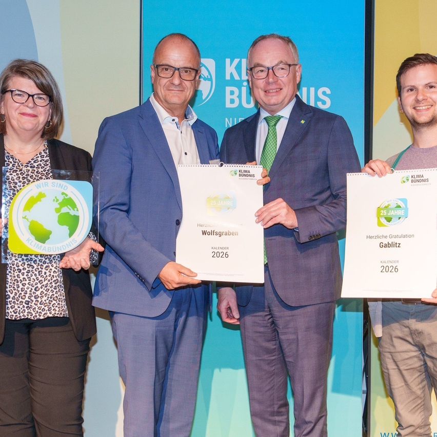 Four individuals stand together holding awards; two men in suits, a woman in a leopard print jacket, and a young man. They are posing for a photo with a banner behind them that says Klimabundnis.