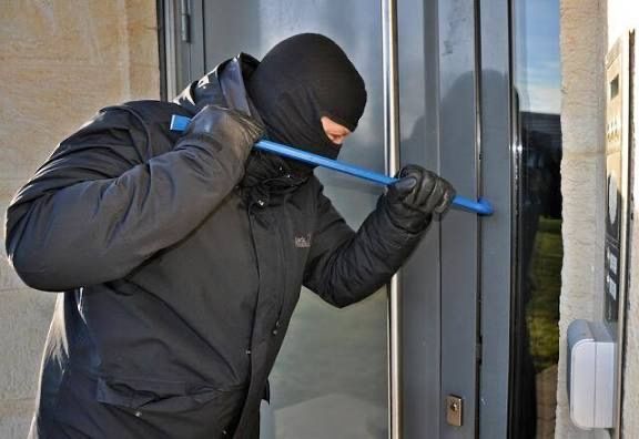 A person wearing a black mask and gloves is holding a blue tool, trying to open a glass door.
