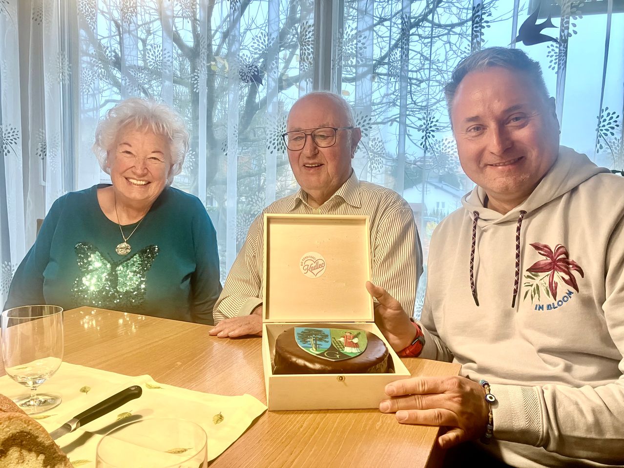 Three individuals seated around a table with a cake in a box, smiling. A woman with white hair and a necklace sits on the left. A man in glasses and a striped shirt sits in the middle. Another man in a white hoodie sits on the right.