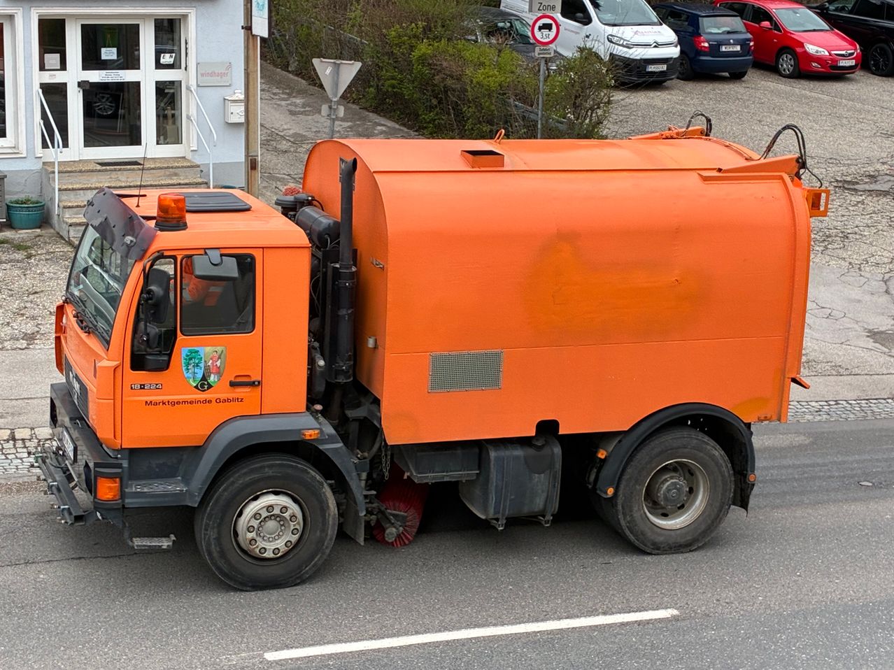 Ein orangefarbener Müllwagen steht auf einer Straße, mit einem Gebäude und Autos dahinter. Der Wagen hat ein Logo an der Seite und ein Schild über dem Fahrerfenster.