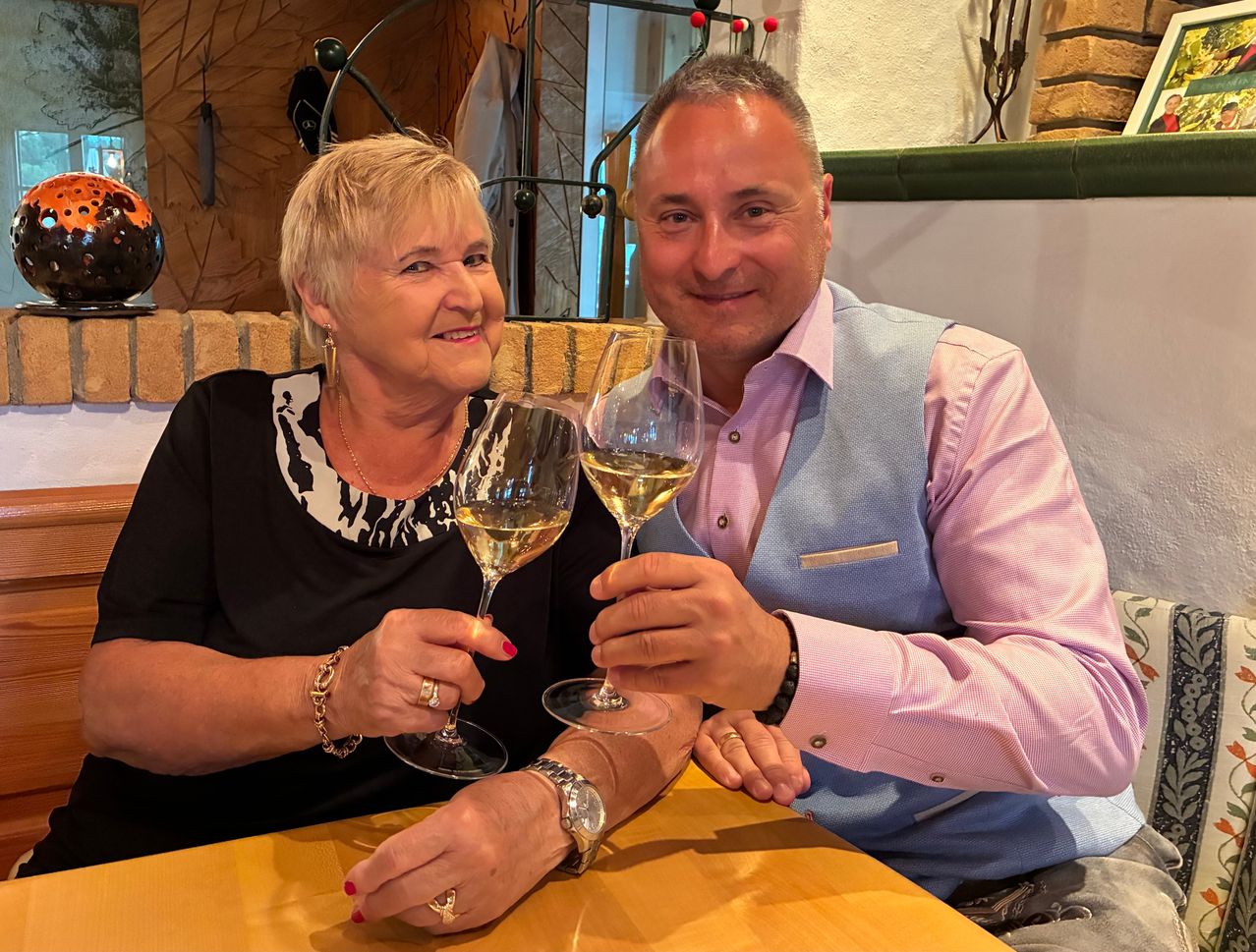A man and woman sit at a table, smiling and clinking wine glasses. The man wears a pink shirt and a blue vest, while the woman wears a black blouse. They both have rings on their fingers.