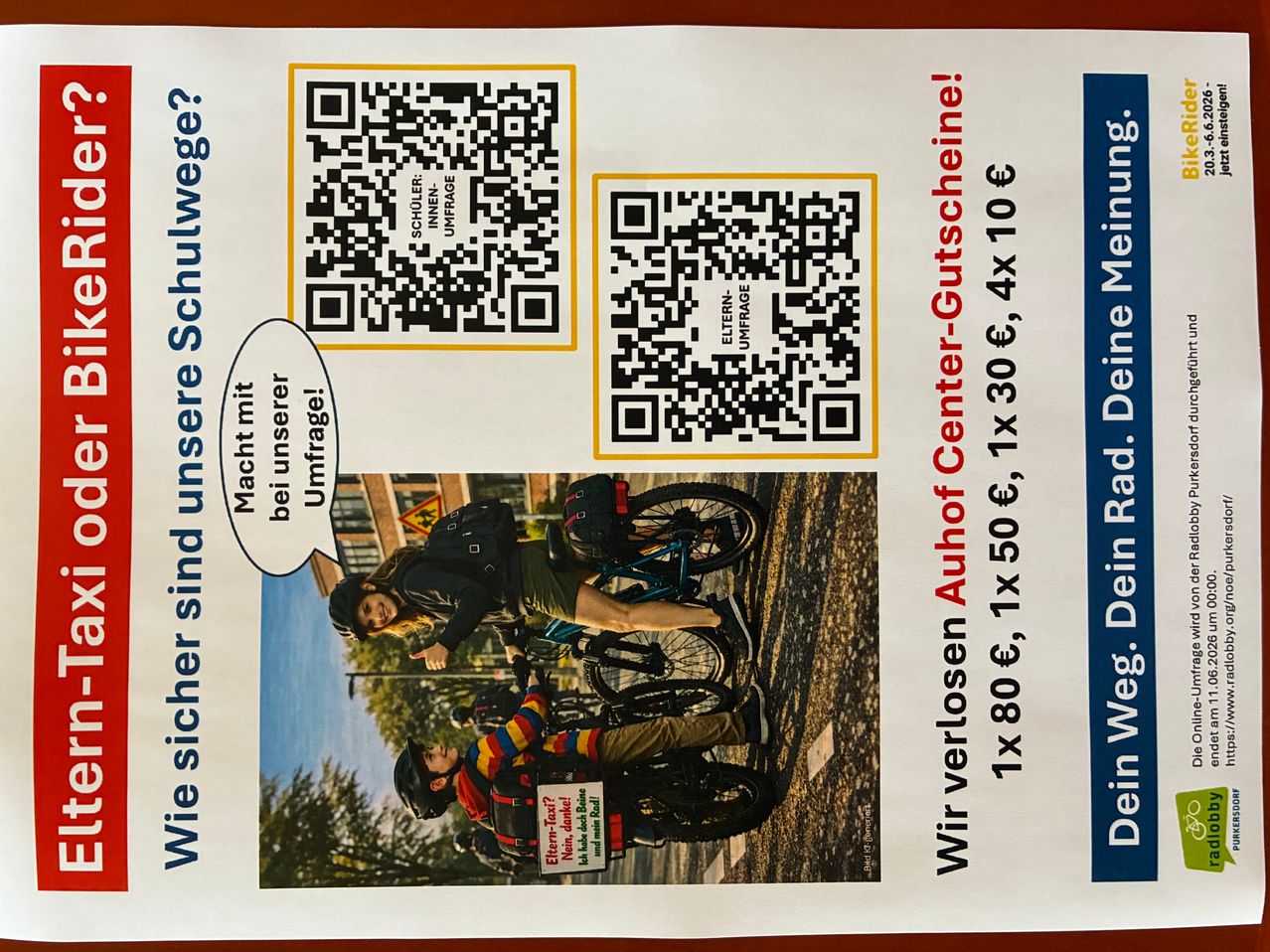 Poster featuring two cyclists and two QR codes. Helmets and backpacks are visible. The text mentions prices for a day, 1x 30€, and 1x 50€. It also says 'Wie sicher sind unsere Schulwege?'.