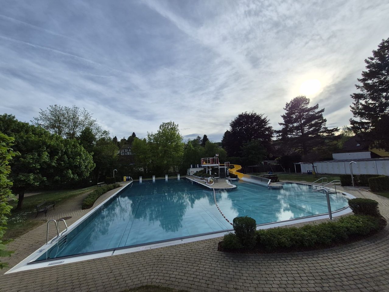 An empty outdoor pool surrounded by green trees and bushes under a cloudy sky with a yellow slide on the right side.