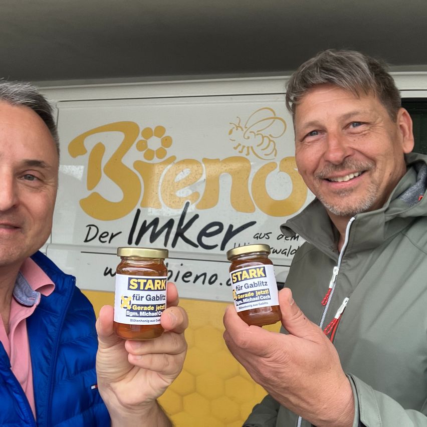 Zwei Männer lächeln und halten Gläser mit Honig mit dem Label 'STARK für Gabitlz' vor einem Fahrzeug mit dem Logo 'Bieno Der Imker'.