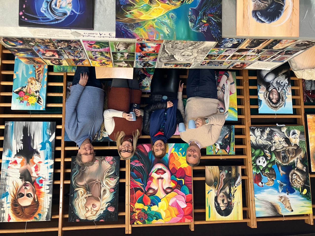 Eine Gruppe von Menschen posiert für ein Foto vor einer Wand voller verschiedener Gemälde. Die Wand ist aus Holz. Sie stehen und sitzen auf dem Boden.