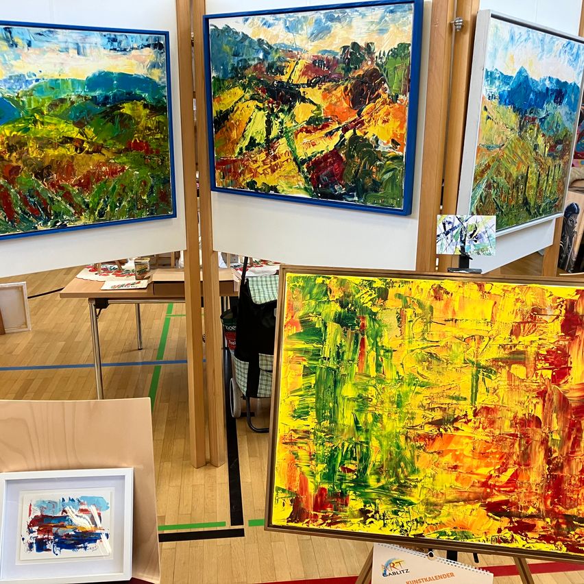 Eine Kunstgalerie zeigt mehrere große Gemälde auf Holzeaseln. Die Gemälde sind abstrakt und haben kräftige Farben. Ein kleines gerahmtes Stück steht auf einem Tisch.