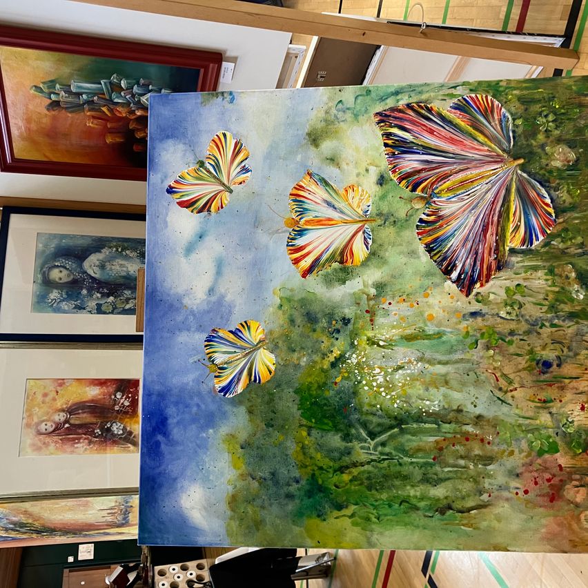 Ein farbenfrohes Aquarellbild zeigt drei bunte Schmetterlinge, die über einer grünen Landschaft fliegen. Der Hintergrund ist blau mit ein paar Wolken. Andere gerahmte Gemälde sind im Hintergrund.
