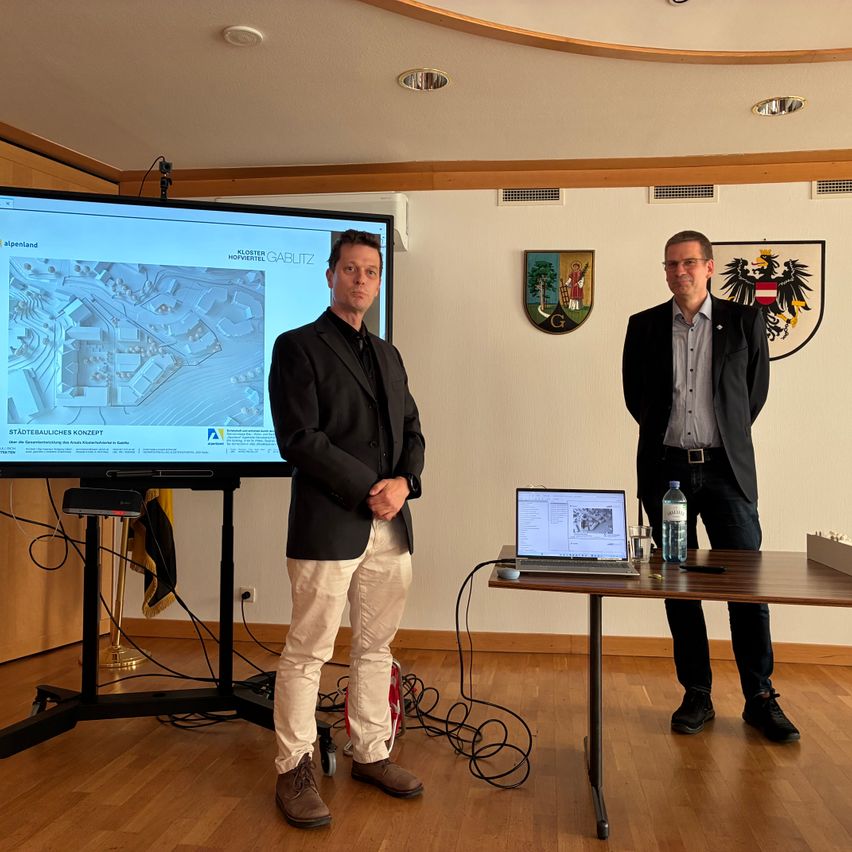 Zwei Männer stehen vor einem Bildschirm mit einer Präsentation. Ein Mann spricht, während der andere auf einem Laptop auf einem Tisch schaut. Auf dem Tisch befinden sich auch Flaschen und eine Schachtel.