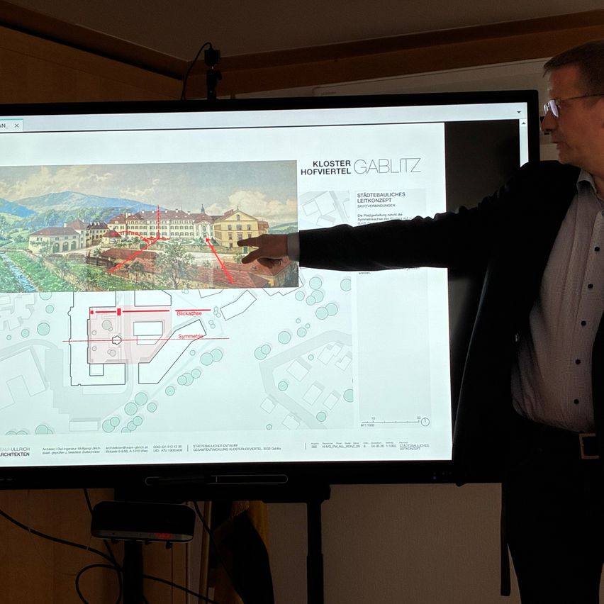 Ein Mann zeigt mit dem Finger auf einen großen Bildschirm, der einen architektonischen Plan von Kloster Hofviertel mit roten Markierungen zeigt. Er trägt einen Anzug und eine Brille.