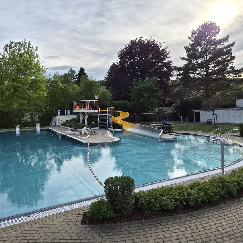 Ein großer Swimmingpool mit einer gelben Wasserrutsche und einer Sprungturm wird von Grünflächen umgeben. Der Pool ist sauber und scheint einsatzbereit zu sein.