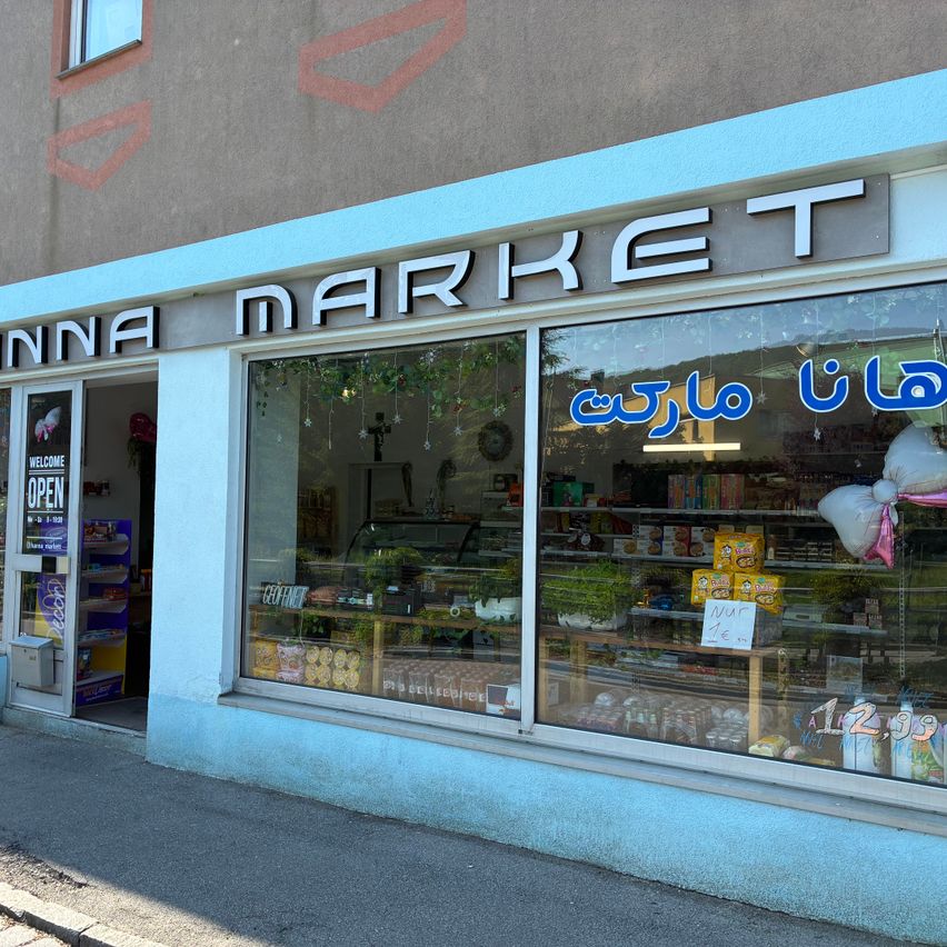 Die Vorderseite eines Geschäfts mit dem Namen Anna Market in großen Buchstaben. Der Laden hat eine Glastür und ein Fenster, das verschiedene Produkte präsentiert. Im Inneren sind Artikel ausgestellt, darunter Pflanzen, Snacks und Luftballons.