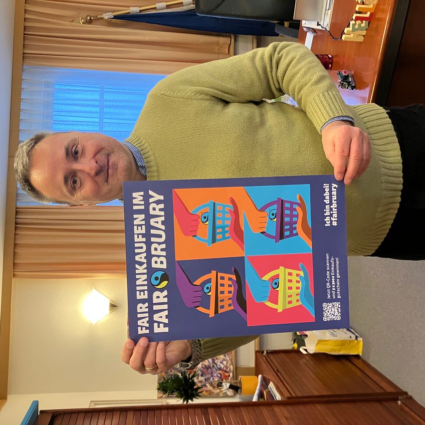A man in a green sweater holds a poster that says 'Fair Einkaufen Im Fair Bruary' in front of a window.