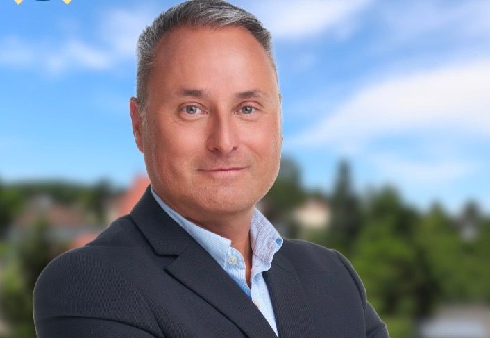 A man in a suit is smiling for the camera with trees and a blue sky in the background.