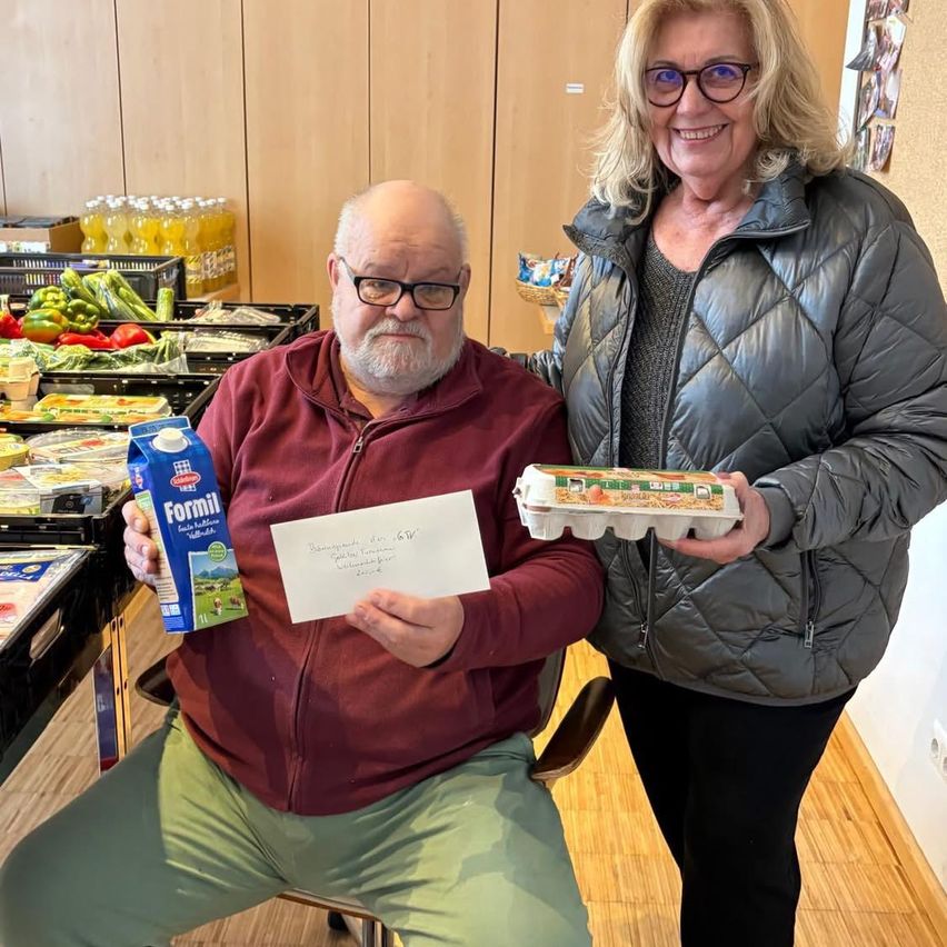 An older man in a maroon hoodie holds a note and a carton of milk while a woman next to him holds a carton of eggs. Behind them is a kitchen with shelves full of bottles and vegetables.