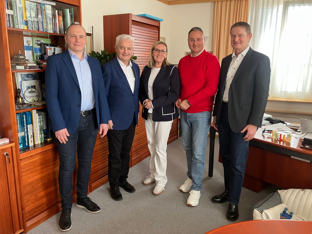 Five people in suits stand in a room with a bookshelf and a desk, smiling for a photo.