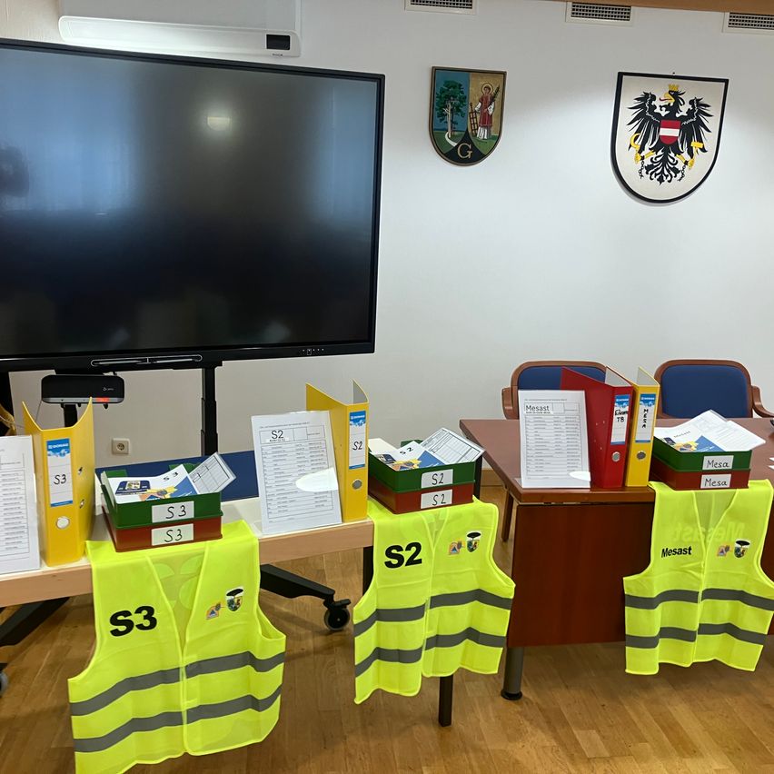 A classroom with a table holding yellow vests labeled S3 and S2, a large monitor, and coats of arms on the wall.