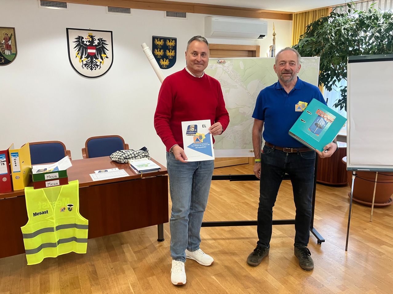 Two men stand in a room with a map and a folder, one wearing a red sweater and the other a blue polo, both smiling. Behind them is a large map on a stand, and various items are on a desk to the left.