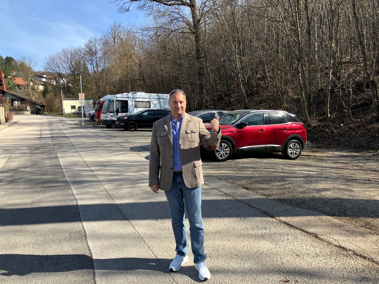 Ein Mann in einem hellbraunen Jackett steht in einem Parkplatz mit geparkten Autos und zeigt den Daumen nach oben. Bäume und ein Gebäude sind im Hintergrund zu sehen.