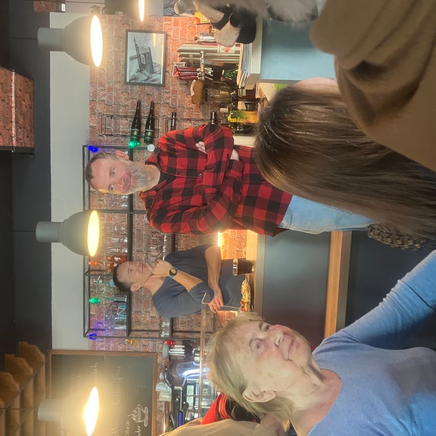 Eine Gruppe von Menschen versammelt sich in einem gemütlichen Restaurant. Ein Mann in einem roten karierten Hemd spricht, während ein anderer Mann zuhört. Zwei Frauen stehen hinter ihnen, eine mit langen Haaren.