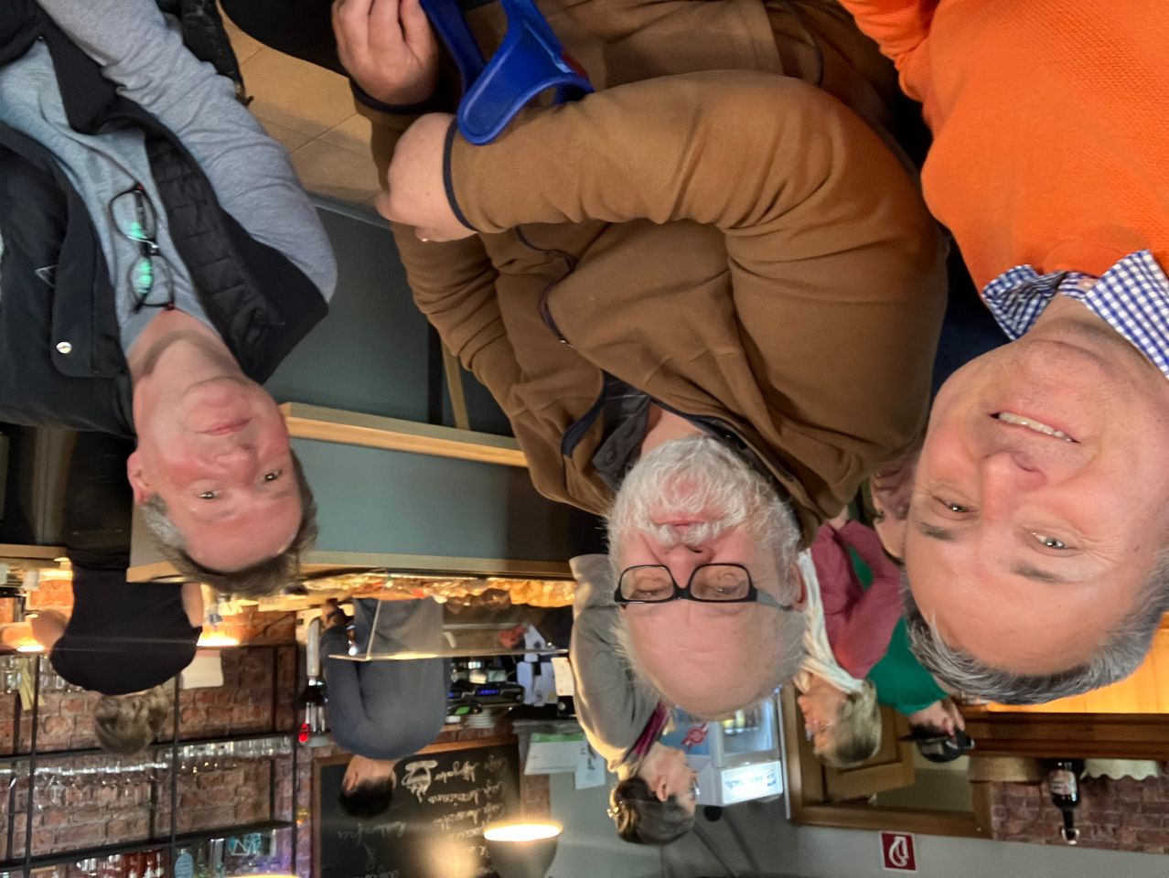 Mehrere Menschen versammeln sich in einer Bar, einer der Männer hängt auf dem Kopf. Der Mann hat einen Bart und eine Brille. Ein anderer Mann schaut von unten zu.