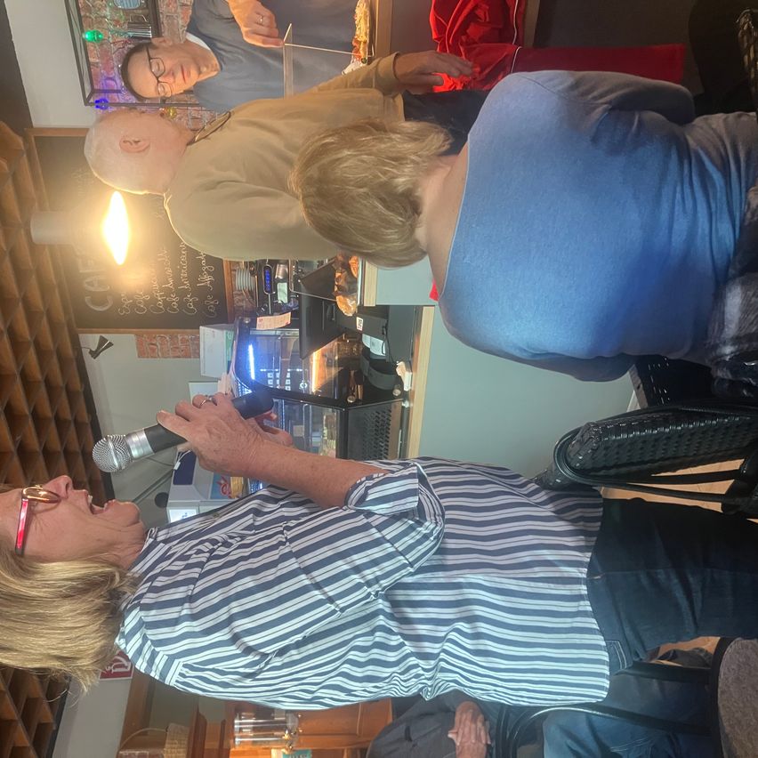 Ein Mann und eine Frau singen in ein Mikrofon in einem Café. Eine weitere Person steht hinter ihnen, und eine Lampe beleuchtet die Szene.