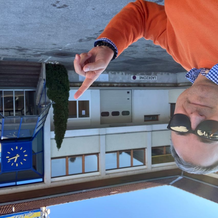 Ein Mann in einem orangefarbenen Pullover zeigt mit dem Finger auf die Kamera vor einem Gebäude mit einer Uhr und Fenstern.