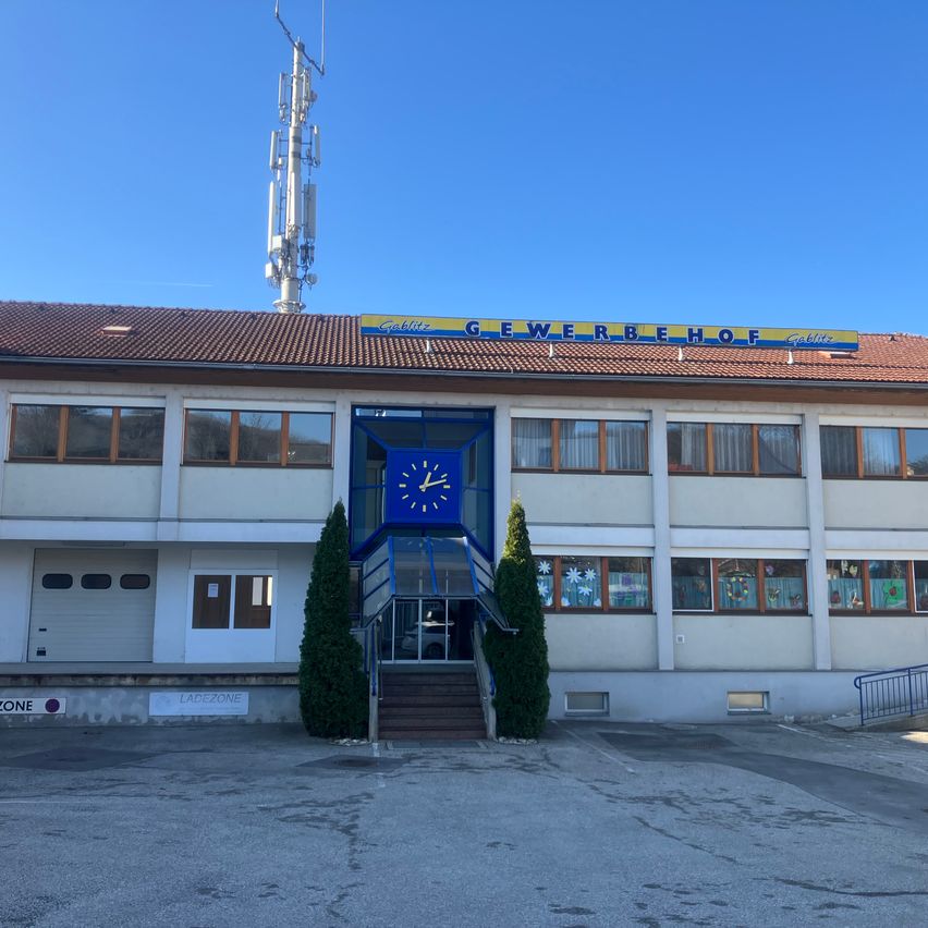 Ein Gewerbegebäude mit einem braunen Dach, einem Turm obenauf und einer blauen Uhr am Eingang. Das Gebäude hat mehrere Fenster und ein kleines Schild an der Seite.