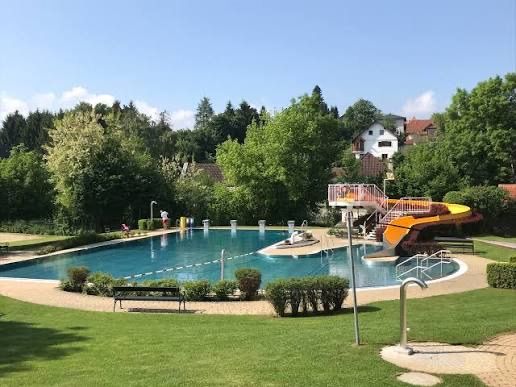 Ein großer Außenpool mit einer gelben Rutsche und einer kleinen Bank in der Nähe eines Grasbereichs. Bäume und Büsche sind in der Umgebung.