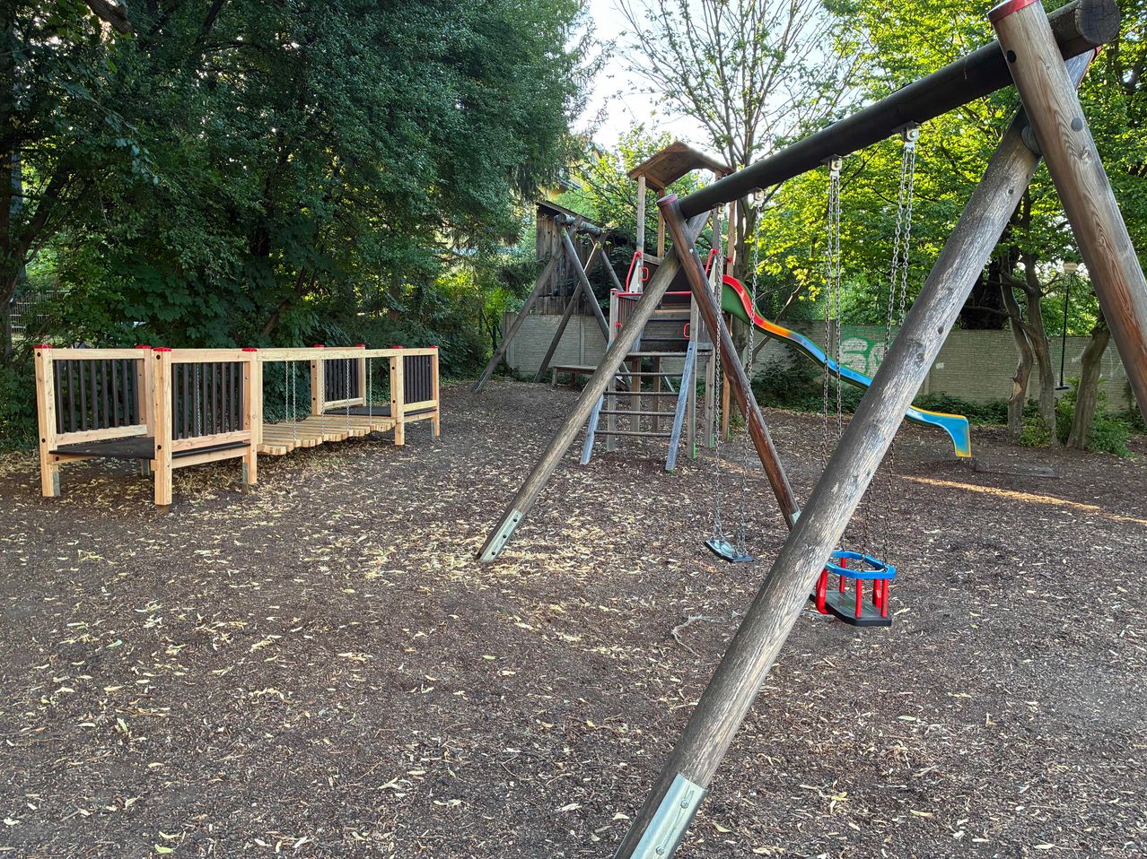 A children's playground with wooden structures, swings, and a slide set in a lush, green environment.