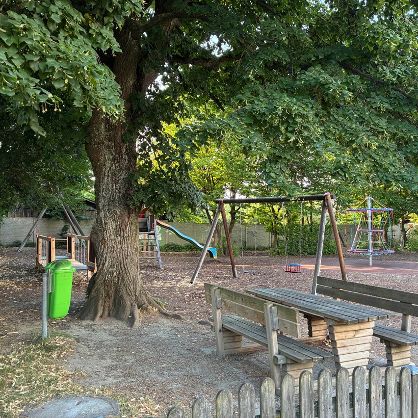 A children's playground with a large tree, a swing set, a slide, and a climbing structure. There are also benches and a green trash bin.