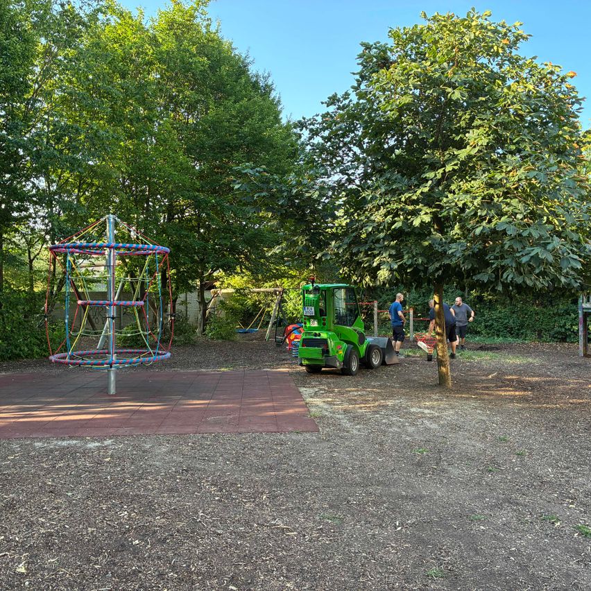 A playground with a colorful climbing structure and a green machinery vehicle parked nearby. People are walking around the area.